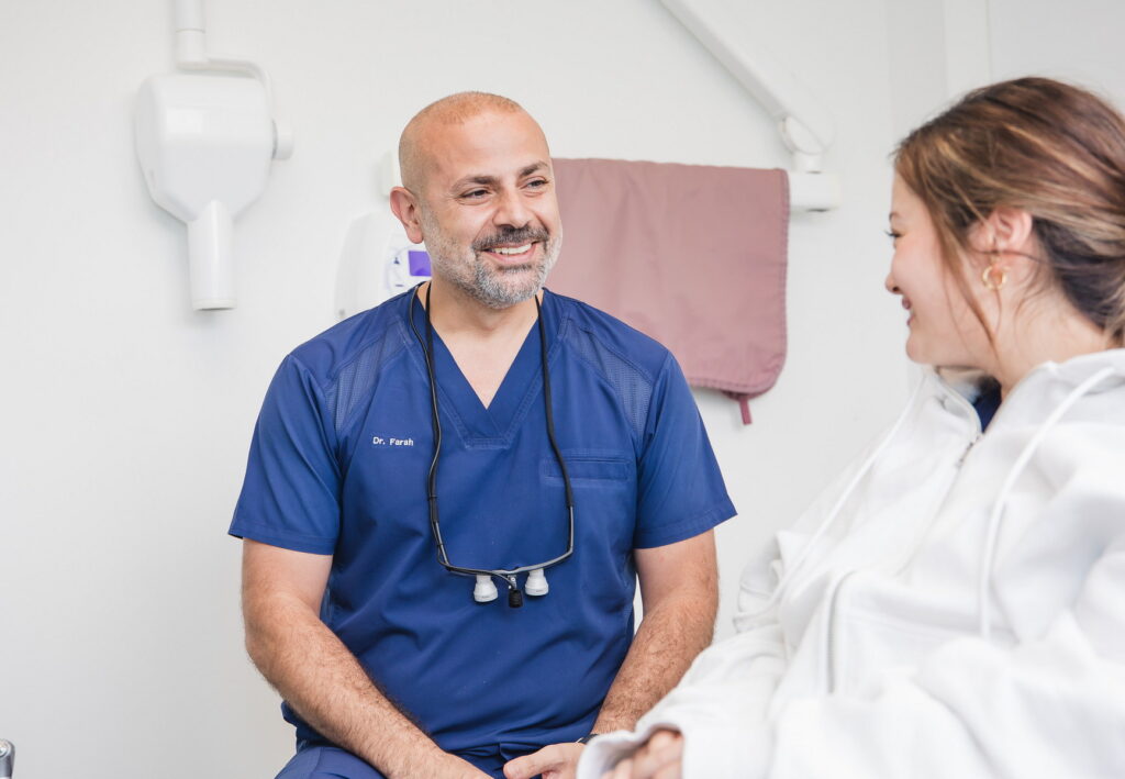 A dentist in blue scrubs smiles while engaging in conversation with a young patient. The setting is a bright dental office, creating a friendly atmosphere.
