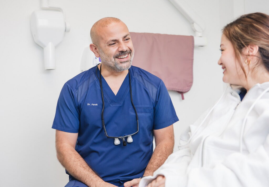 A friendly dentist in blue scrubs smiles at a patient during a consultation in a bright dental office. The patient, wearing a white hoodie, appears relaxed and engaged in conversation, creating a welcoming atmosphere for dental care.