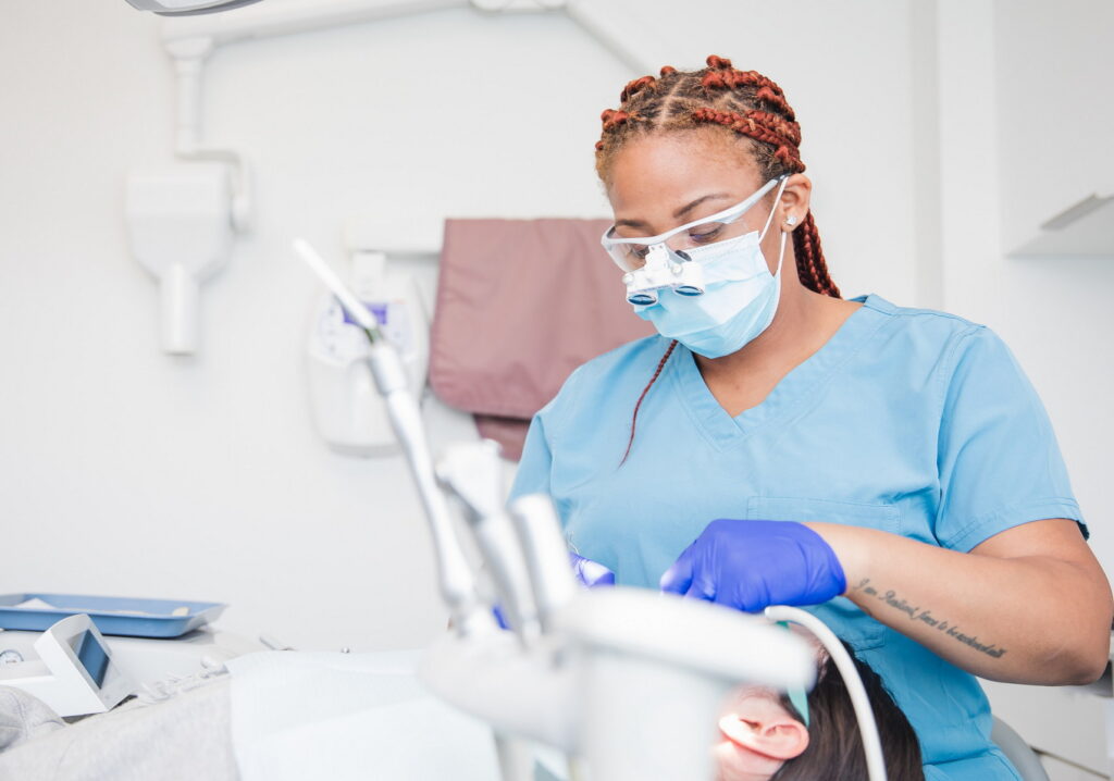 A dental professional with braided hair, wearing glasses and a face mask, is attentively working on a patient's mouth in a clean dental office environment.