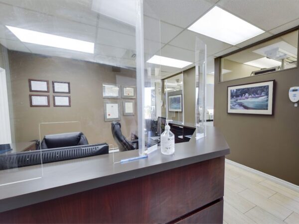 Dental office reception area with a counter, protective screens, and hand sanitizer. Comfortable seating and framed artwork are visible, along with bright lighting and large windows.
