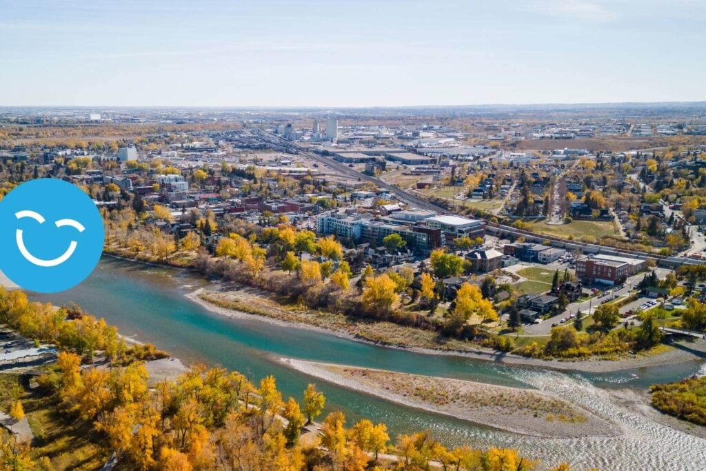 An aerial view of a vibrant riverside city in autumn, showcasing colorful trees along the riverbank and urban development in the background. A blue smiley icon is positioned in the corner, adding a cheerful touch to the scenery.
