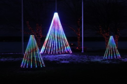 Colorful light displays shaped like Christmas trees are arranged in a dark outdoor setting, with bright neon lights creating a festive atmosphere. The background features a river and trees adorned with additional lights.