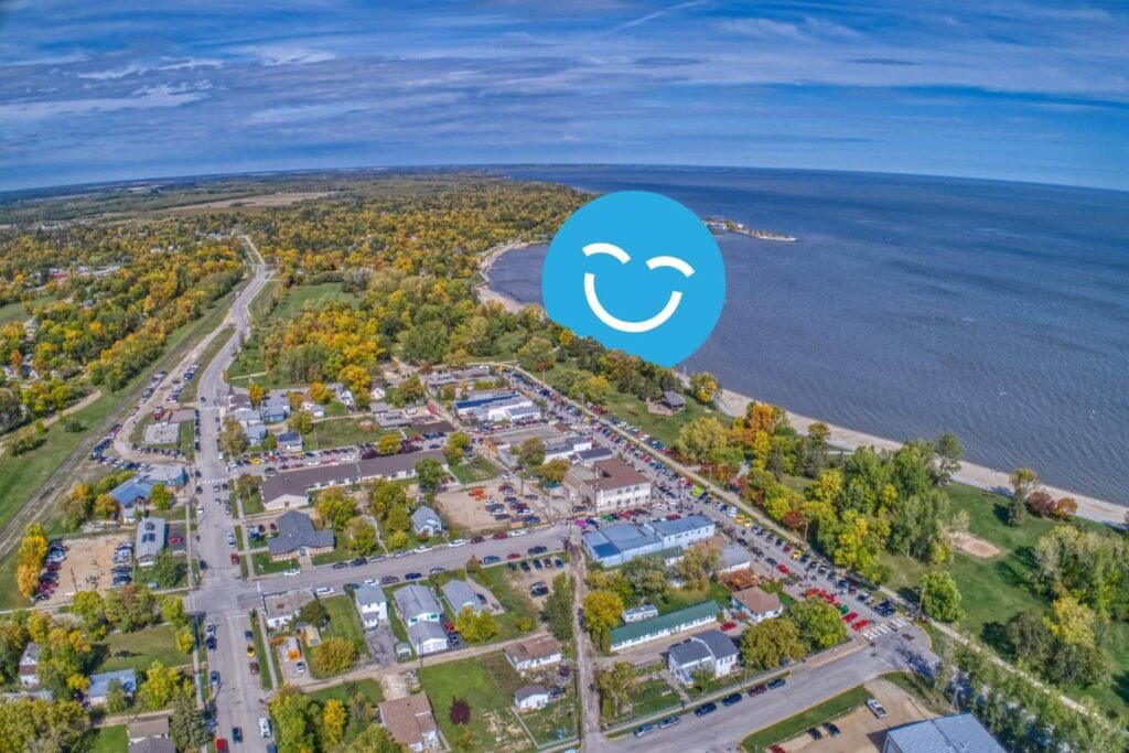 Aerial view of a small town by the water, surrounded by trees featuring autumn colors. A smiling face icon overlays the image, adding a cheerful element to the landscape.