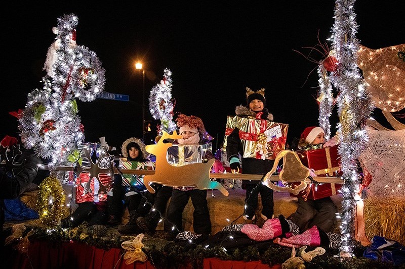 A festive parade float featuring performers dressed in colorful costumes, surrounded by holiday decorations and lights. The scene is set at night, creating a vibrant and cheerful atmosphere.