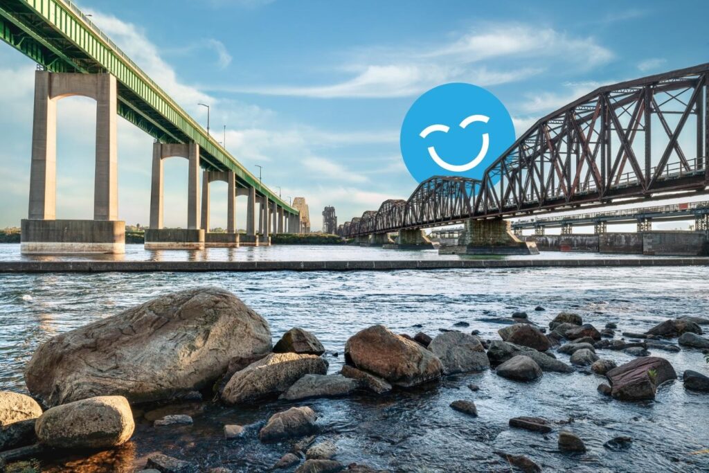 A scenic view of a river featuring two bridges: a green highway bridge and a brown railroad bridge. Smooth rocks line the water's edge under a bright sky, with a cheerful blue smiley face icon overlaying the image.