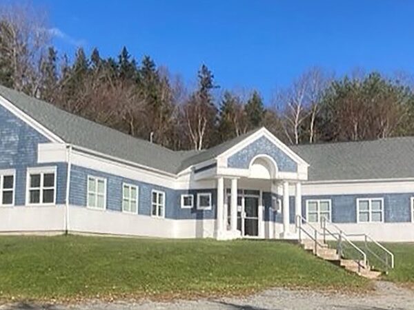A modern, single-story building features a blue and white exterior with large windows and a centered entrance. It is surrounded by green grass and trees, set against a clear blue sky.