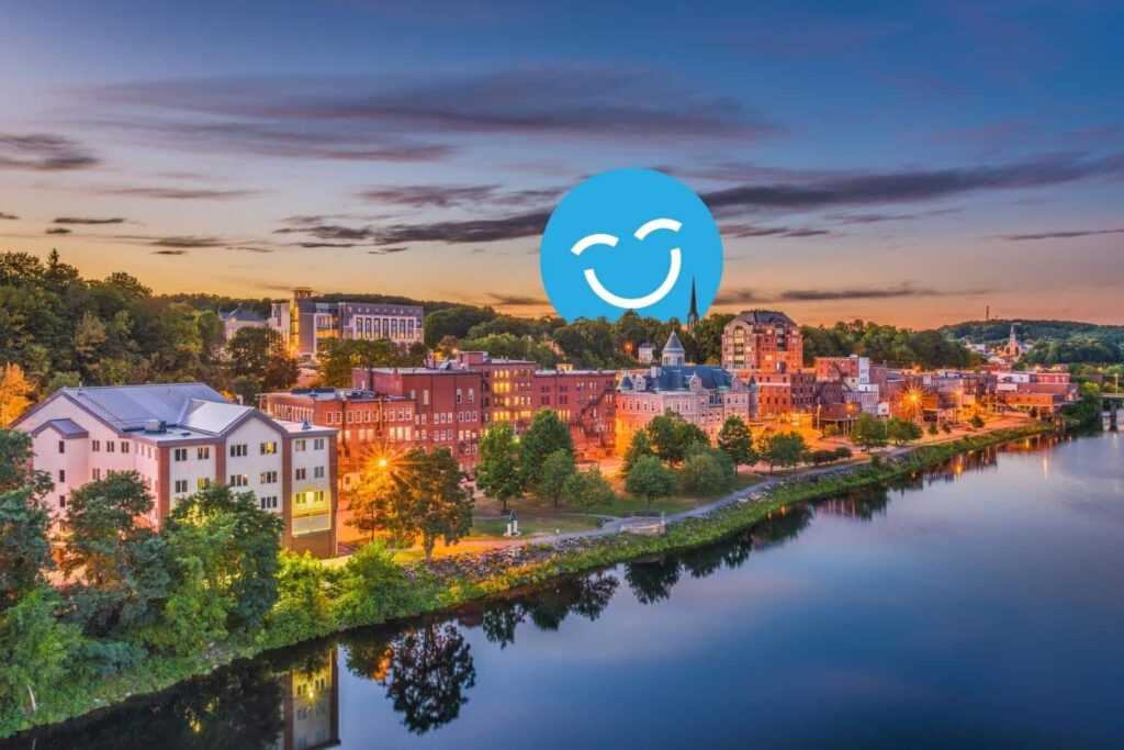 A picturesque view of a riverside town at sunset, featuring charming buildings along the water with trees and reflections. A blue smiley face icon overlays the image, adding a cheerful element to the scene.