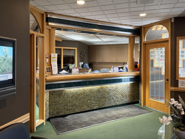 A welcoming dental office reception area featuring a wooden counter, comfortable seating, and a television. Natural light streams through a glass door, creating an inviting atmosphere. The 123Dentist Smiley adds a friendly touch to the environment.