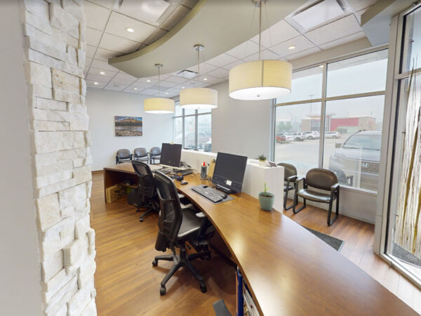 A modern dental office featuring a reception area with sleek wooden desks, computer stations, and comfortable chairs. The space is bright and inviting, with natural light streaming through large windows and stylish pendant lighting hanging overhead. The 123Dentist Smiley adds a friendly touch to the environment.
