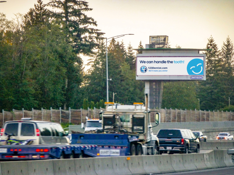 A busy highway scene featuring various vehicles in traffic, with a roadside billboard displaying the message "We can handle the tooth" alongside the friendly 123Dentist Smiley. Trees line the background, indicating a natural setting.