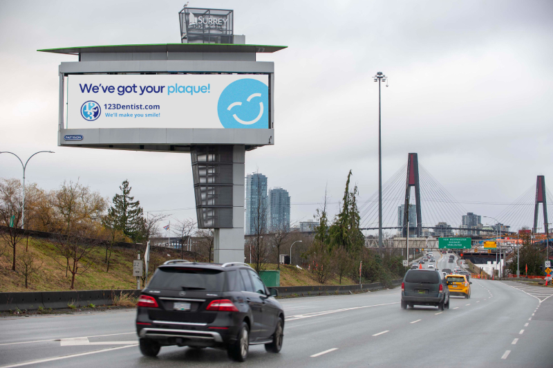 A digital billboard on a busy highway promotes dental health with the message "We've got your plaque!" featuring the friendly 123Dentist Smiley. Cars are seen driving past the billboard, which is surrounded by a cityscape and trees.