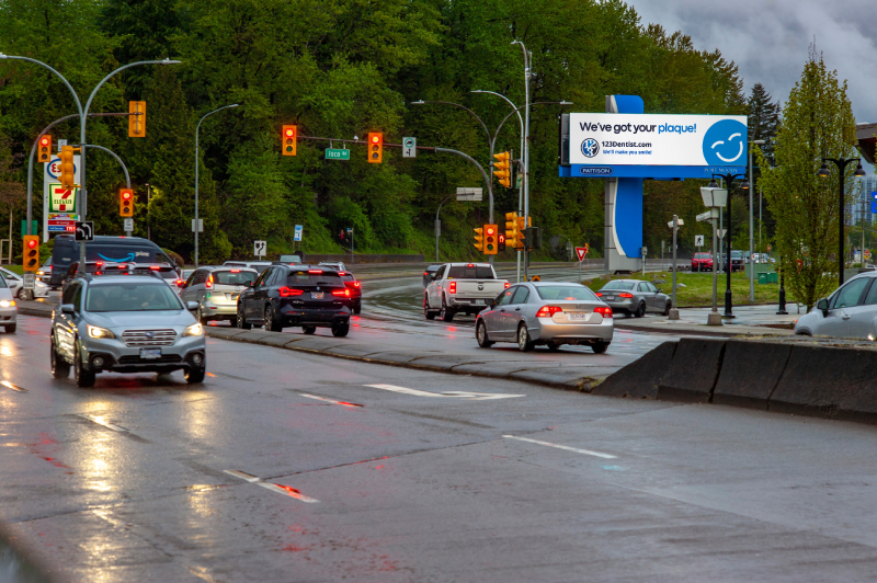 A busy urban street scene with vehicles stopped at traffic lights. A large billboard featuring the 123Dentist Smiley advertises dental services, surrounded by greenery and overcast skies.