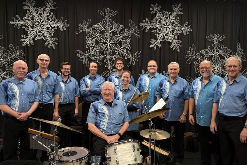 A group of twelve musicians wearing matching blue shirts pose for a photo, with one member seated at a drum set. The background features decorative snowflakes, creating a festive atmosphere.