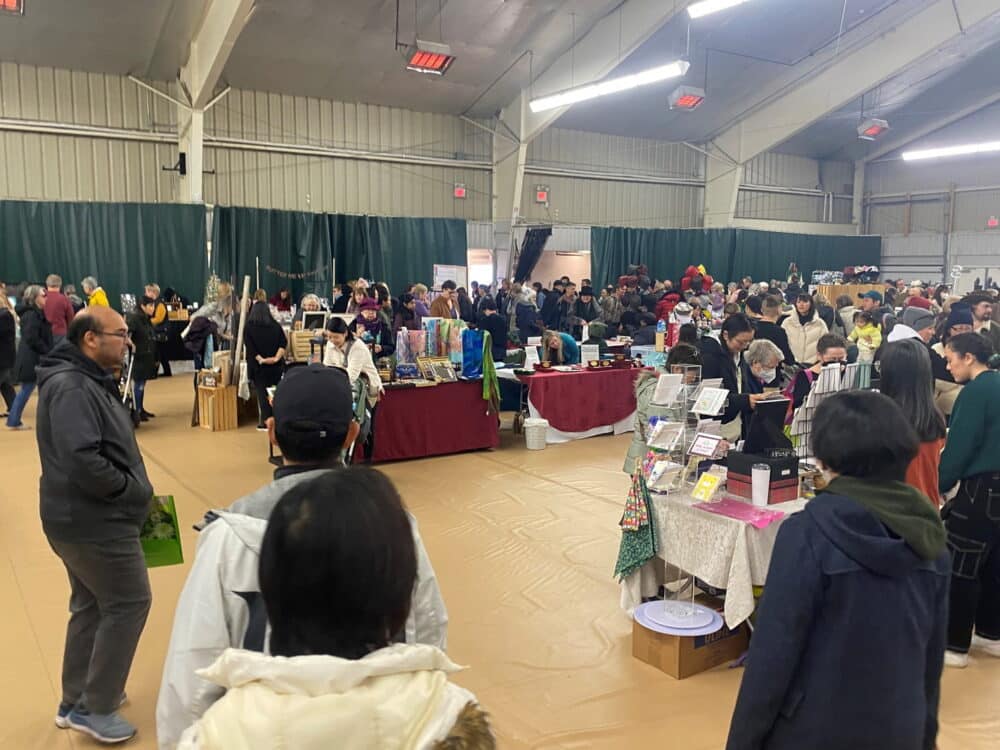 A bustling indoor market scene filled with visitors browsing various stalls. Tables are lined with products, while attendees engage with vendors. The space is lively, highlighting community interaction in a vibrant setting.
