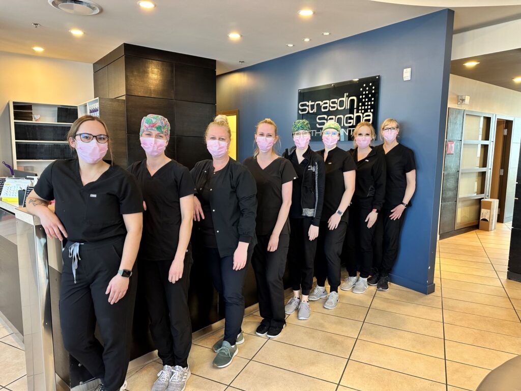 A group of eight dental professionals stands together in a dental office, all wearing black scrubs and face masks. They smile warmly, promoting a friendly environment. The backdrop features a sign that reads "Strength in Smiles." The atmosphere is welcoming and professional.