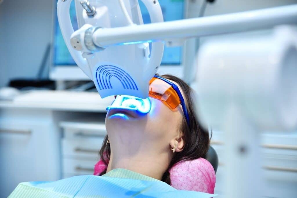 A woman sits in a dental chair, wearing protective glasses, while a dental light illuminates her mouth. The setting suggests a dental examination or treatment, with equipment visible in the background. The mood is calm and professional, emphasizing dental care.