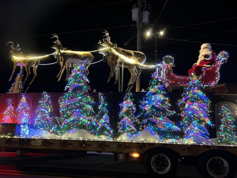 A festive holiday parade float features Santa Claus riding in a sleigh pulled by reindeer, surrounded by sparkling, multi-colored Christmas trees adorned with twinkling lights. The scene is illuminated against a dark night sky, creating a warm, cheerful atmosphere.