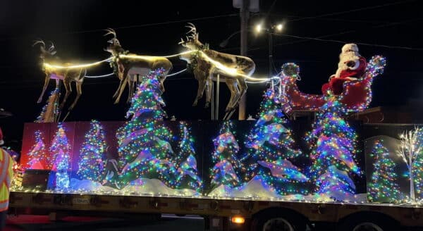 A festive holiday parade float features Santa Claus riding in a sleigh pulled by reindeer, surrounded by sparkling, multi-colored Christmas trees adorned with twinkling lights. The scene is illuminated against a dark night sky, creating a warm, cheerful atmosphere.