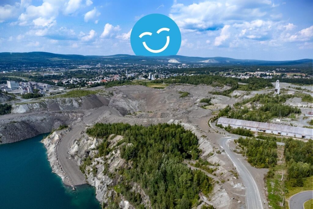 Aerial view of a large quarry surrounded by greenery, with a small lake nearby. A city skyline and hills are visible in the background. A blue smiley face emoji is overlaid on the sky.
