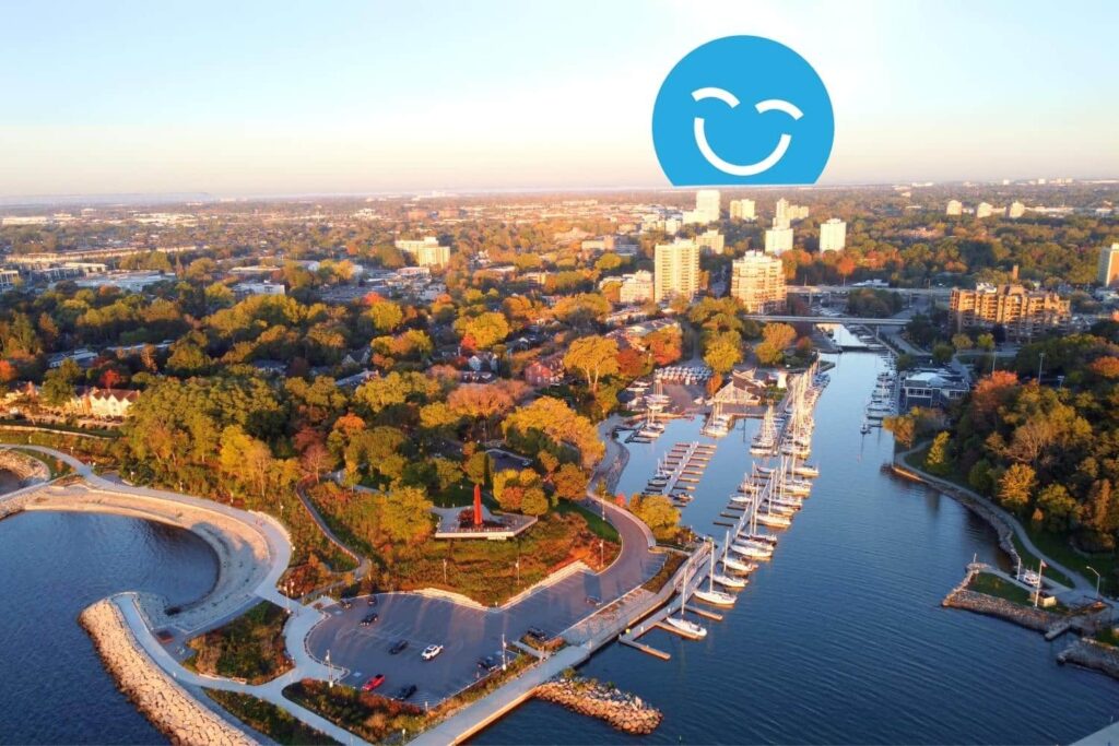 Aerial view of a coastal city during sunset, featuring a marina with docked boats, lush greenery, and cityscape in the background. An overlaid smiling blue face icon is at the top.