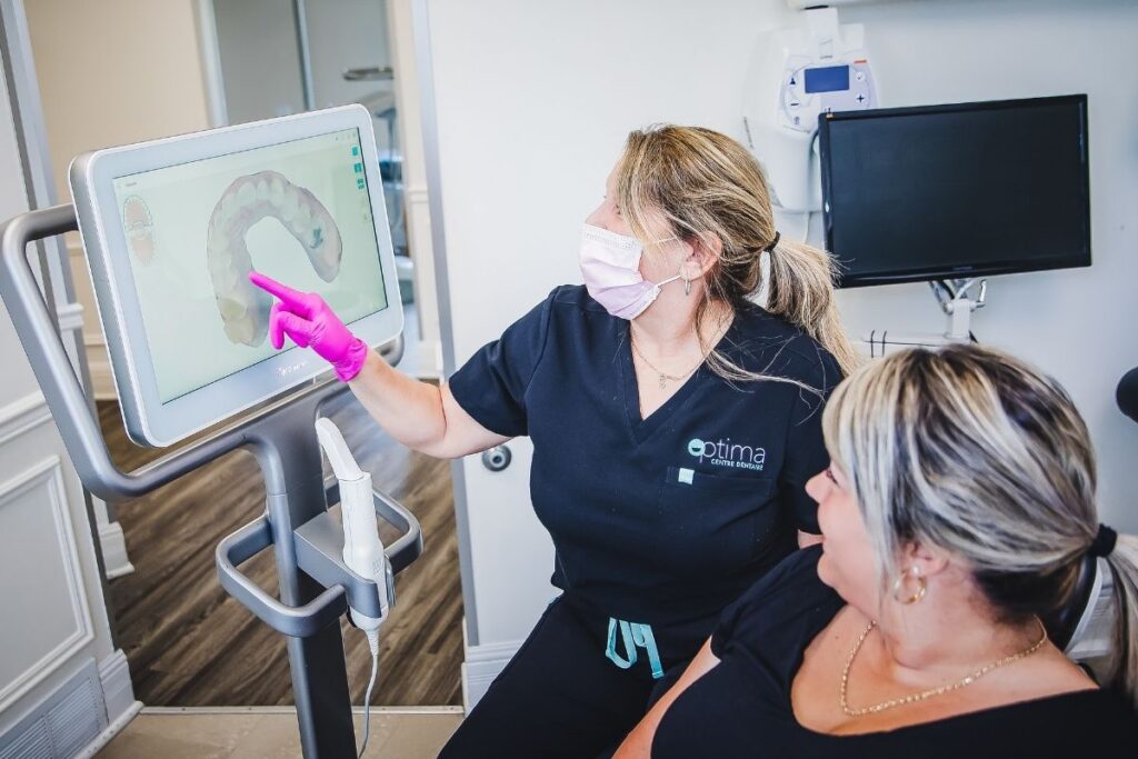 A dental professional in gloves points at an interactive screen while explaining something to a patient seated nearby. Both individuals are wearing masks in a bright, modern dental office setting.