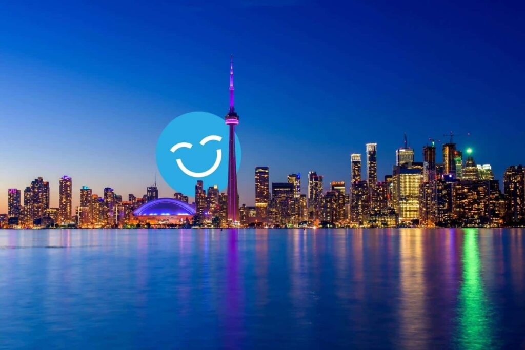 Toronto skyline at dusk, featuring illuminated skyscrapers and the CN Tower reflecting on Lake Ontario. A smiley face icon overlays part of the scene.
