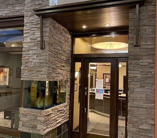 A dental office entrance featuring a modern stone facade, glass doors, and a lighted sign reading "towne DENTAL." The lobby is visible through the doors, and patterned tile flooring leads up to the entrance.