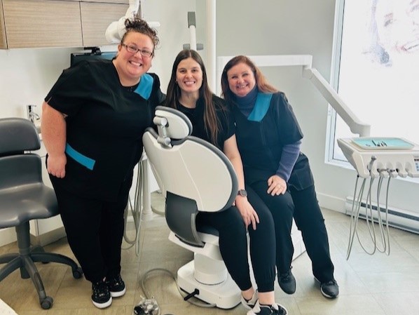 Three dental professionals, wearing matching black scrubs with blue accents, stand together smiling in a dental office. A dental chair is positioned in front of them, with dental tools visible nearby. The friendly 123Dentist Smiley adds a cheerful touch to the scene.