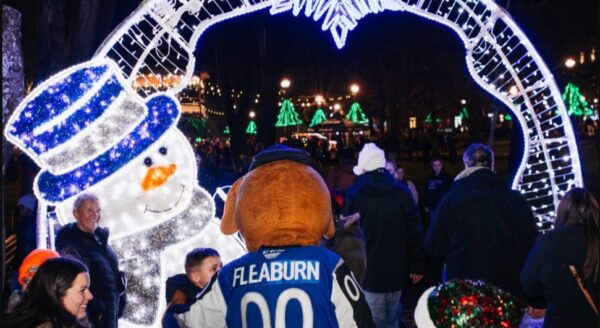 A festive scene featuring a large illuminated snowman and an archway decorated with lights. A person dressed in a mascot costume stands among a crowd, with holiday decorations in the background, creating a cheerful atmosphere.