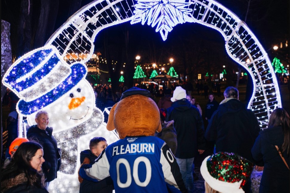 A festive scene featuring a large illuminated snowman and an archway decorated with lights. A person dressed in a mascot costume stands among a crowd, with holiday decorations in the background, creating a cheerful atmosphere.