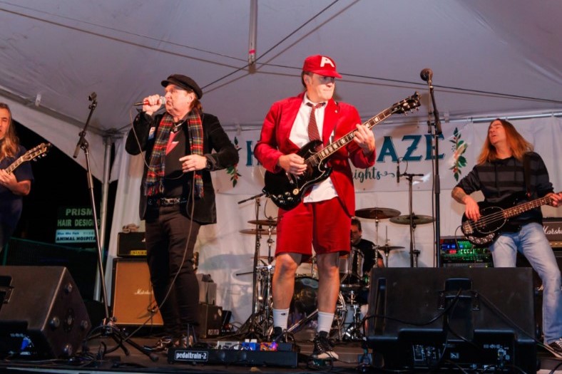 A band performs on stage under a tent, featuring a lead singer with a scarf, a guitarist in a red outfit, and other musicians. Musical equipment is set up behind them, creating a lively atmosphere.
