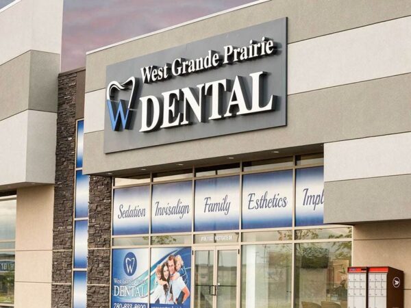 A modern dental clinic facade featuring a prominent sign that reads "West Grande Prairie Dental." The entrance showcases large windows displaying various dental services, creating an inviting and professional atmosphere.