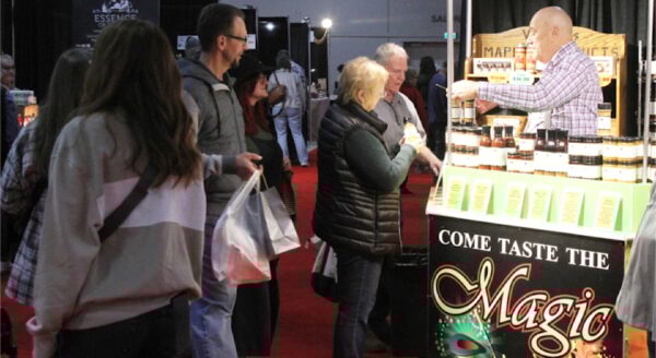 A lively market scene shows a vendor offering samples at a booth labeled "Come Taste the Magic." Customers engage with the vendor while exploring various products on display, surrounded by other stalls in a bustling event space with a vibrant red carpet.