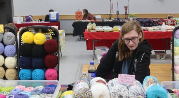 A person sits at a table filled with colorful yarn skeins, surrounded by additional vendor booths in a market setting. The atmosphere is vibrant and inviting, showcasing a variety of crafts and handmade goods.