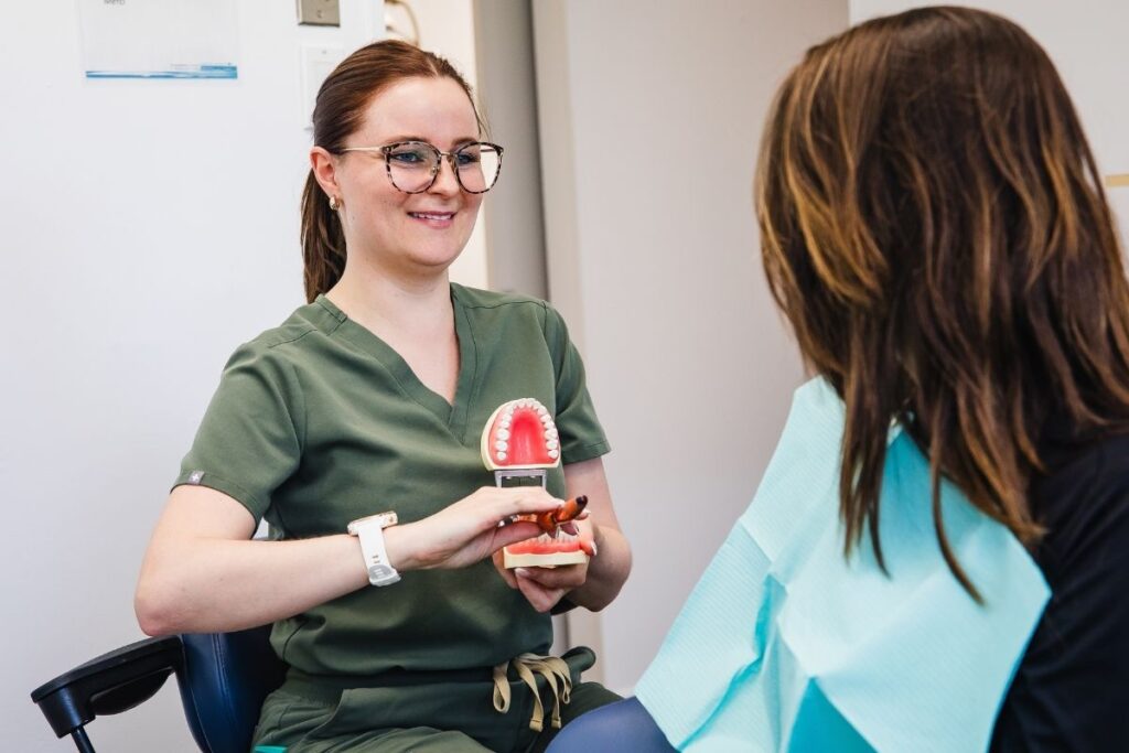 A dental professional in green scrubs smiles while showing a dental model to a patient seated in a dental chair. The patient is watching attentively, draped in a blue dental bib.