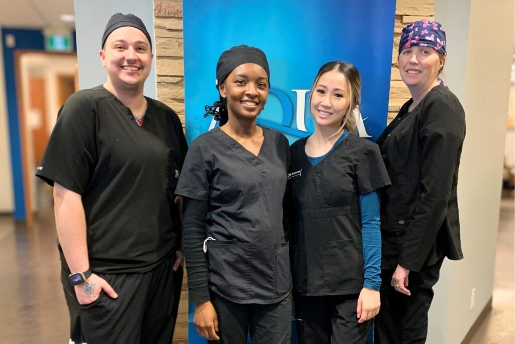 A group of four dental professionals, wearing black scrubs, smile confidently in a clinic setting. They stand in front of a blue backdrop featuring the 123Dentist logo. The atmosphere is friendly and welcoming, showcasing teamwork and professionalism.