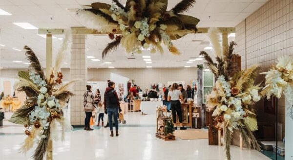An event space featuring a floral arch made of dried plants and flowers, with people browsing stalls in the background. The setting is bright and spacious, indicating a gathering or market.