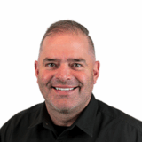 A smiling man with short gray hair and a beard, wearing a black button-up shirt, is depicted against a white background.