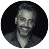 A smiling man with silver hair, dressed in a dark shirt, poses in a circular frame against a black background, exuding warmth and friendliness.