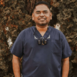 A smiling man in a dark medical uniform stands in front of a textured, brown backdrop, wearing dental loupes around his neck.