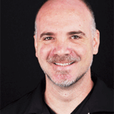 A smiling man with short, gray hair and a beard is wearing a black shirt. The background is plain black, highlighting his facial features and expression.