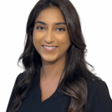 A smiling healthcare professional wearing dark scrubs, with long dark hair styled in waves. The background is plain white, highlighting the individual's friendly expression and professional attire.