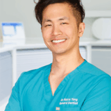 A smiling male dentist wearing a turquoise scrub top stands with his arms crossed, showcasing a confident and approachable demeanor in a dental office setting.