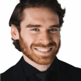 A smiling man with a beard and wavy hair is dressed in a formal black suit and tie, against a plain white background.