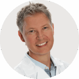 A smiling male doctor with short gray hair, wearing a white lab coat over a light blue checkered shirt, is pictured against a plain background.